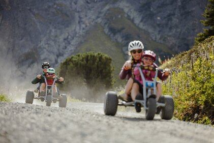 Two family members ride downhill in separate mountaincarts on a dusty gravel path. On the right, a woman rides with a child in a mountaincart, the child seated in front. On the left, a man rides with a child in the front mountaincart, surrounded by kicked-up dust. Mountain scenery and green vegetation are visible in the background | © Gargellner Bergbahnen GmbH & Co KG, Alexander Fuchs 