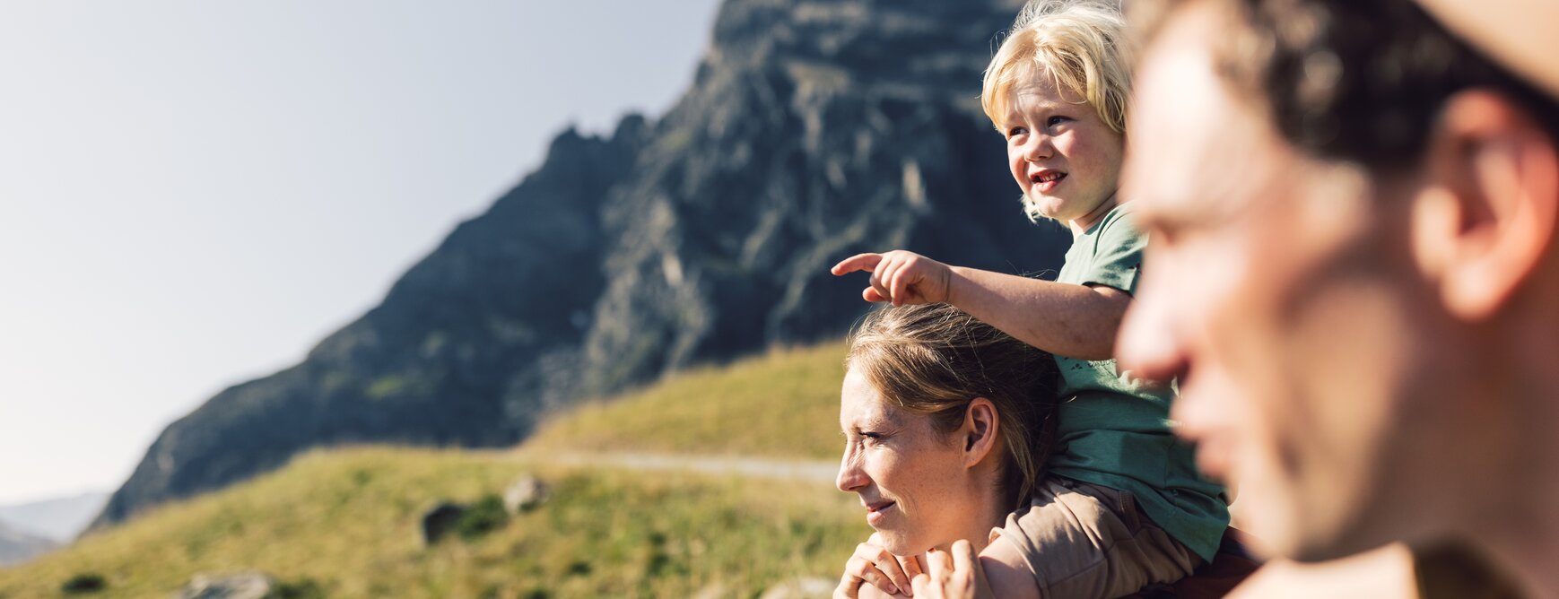 Ein Mann und eine Frau stehen auf einer blühenden Bergwiese in Gargellen im Montafon. Auf den Schultern der Frau sitzt ein blonder Junge, der mit dem Finger neugierig in die Ferne zeigt. Gemeinsam blicken sie in die Weite – im Hintergrund erhebt sich die imposante Madrisa unter einem strahlend blauen Himmel. Die Szene ist in einer Nahaufnahme festgehalten. | © Gargellner Bergbahnen GmbH & Co KG, Daniel Zangerl