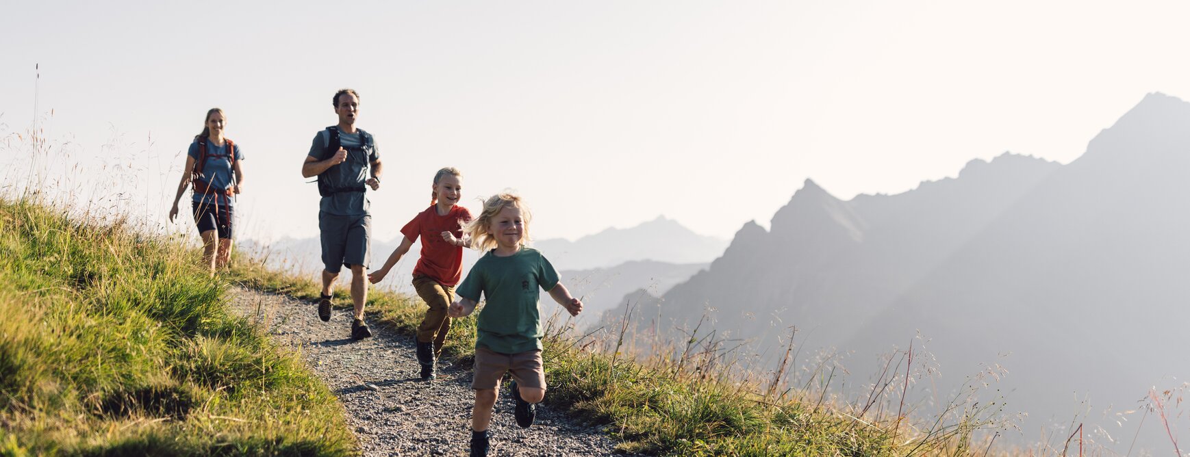  A family with two adult hikers and two children is laughing and running up a gravel path in Gargellen in Montafon region. The children, a boy with blonde hair in a green t-shirt and a girl in a red t-shirt, are joyfully running ahead. Gentle mountain slopes are visible in the background under a bright sky. | © Gargellner Bergbahnen GmbH & Co KG, Daniel Zangerl