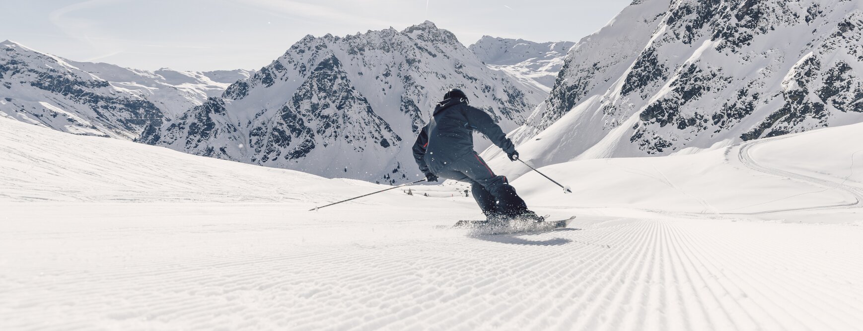 A skier is seen from behind, carving down the freshly groomed slope as snow sprays up around them. In the background, the Madrisa and the snow-covered Ritzenspitzen peaks rise into the sky. | © Gargellner Bergbahnen GmbH & Co KG, Daniel Zangerl